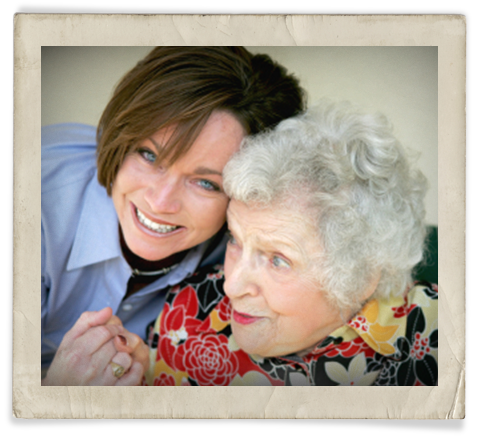 A young, blue-eyed caregiver hugs an older, white-haired woman.